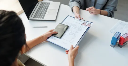 Two people sit at a desk; one hands a clipboard with a form, photos, and passport to the other. A laptop, pen, and paper trays are on the table—an everyday scene for a Chicago employment immigration lawyer.