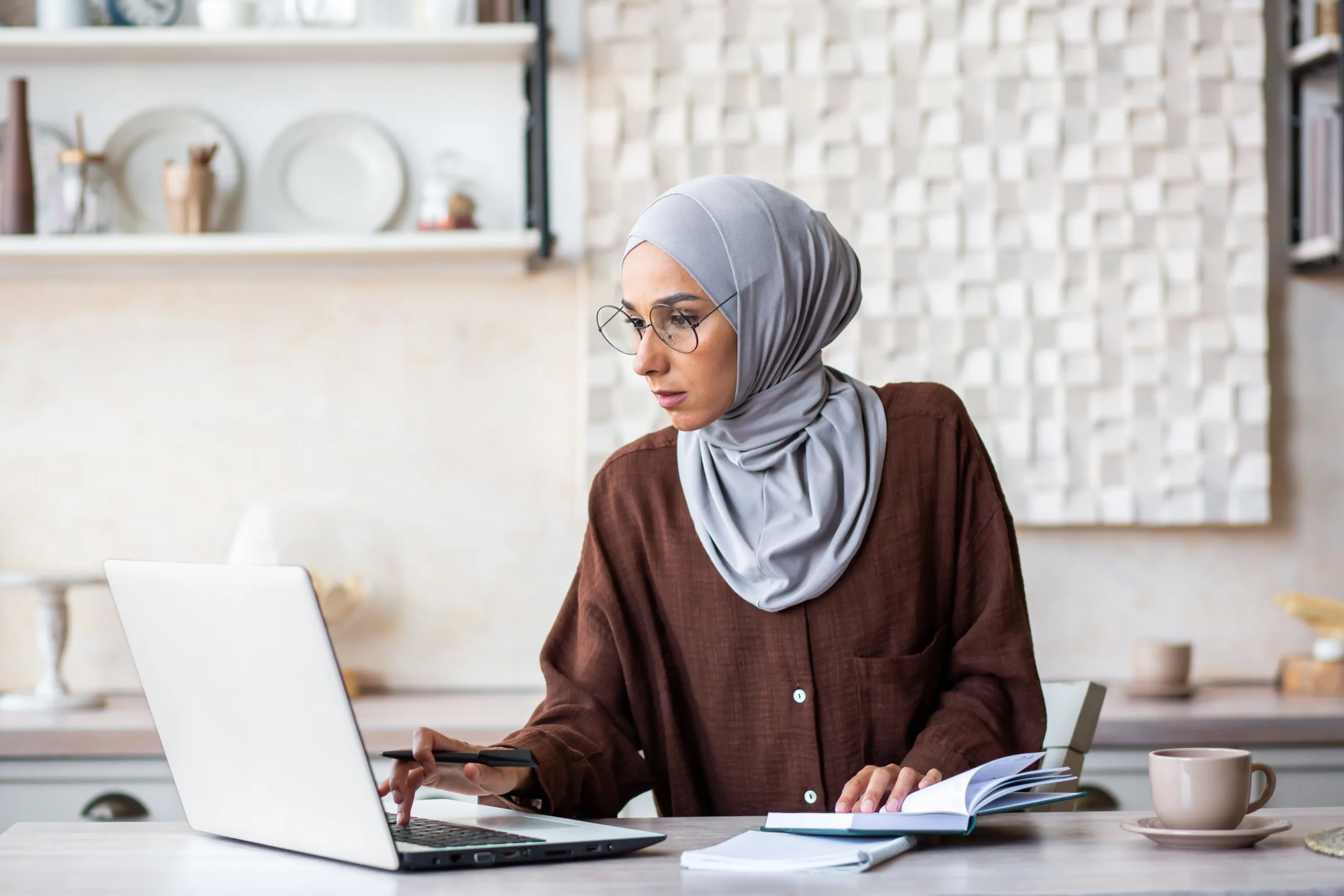 Woman wearing a gray hijab and glasses works at her laptop, pen in hand, with an open notebook beside her—she might be researching Chicago immigration attorneys in the comfort of her modern kitchen.