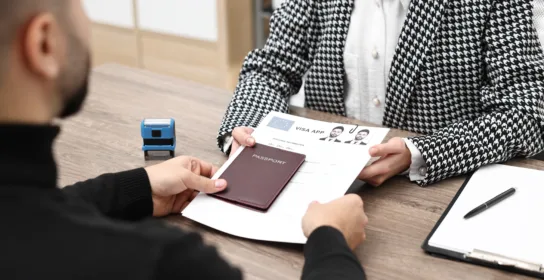 Person hands over a passport and visa application form to an official sitting at a desk with a stamp and clipboard, seeking guidance from an experienced employment visa lawyer in Chicago.